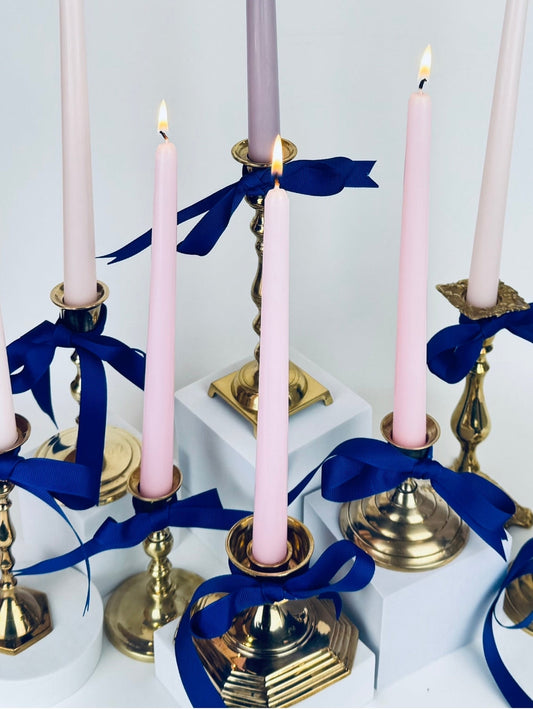 Assorted vintage brass candlestick holders arranged in a cluster, each topped with pastel taper candles and tied with deep blue ribbon, styled on a white surface with soft shadows.