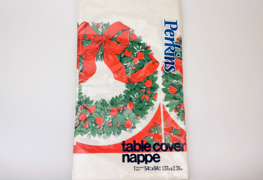 Factory-sealed vintage Perkins Christmas table cover featuring a bold red bow and green wreath print on a white background, with the blue Perkins logo and size label 54 in × 94 in visible on the packaging.