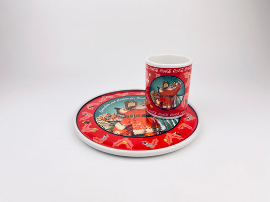 Vintage 1998 Coca-Cola Santa cookie plate and matching cup by Enesco, featuring Santa Claus drinking Coca-Cola with red holiday graphics and holly border, displayed on a white background.