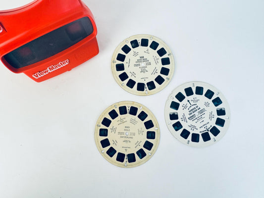 Vintage red View-Master with three stereoscopic reels featuring scenes from Florida USA, Basle Switzerland, and Garden of the Gods, displayed on a white surface.