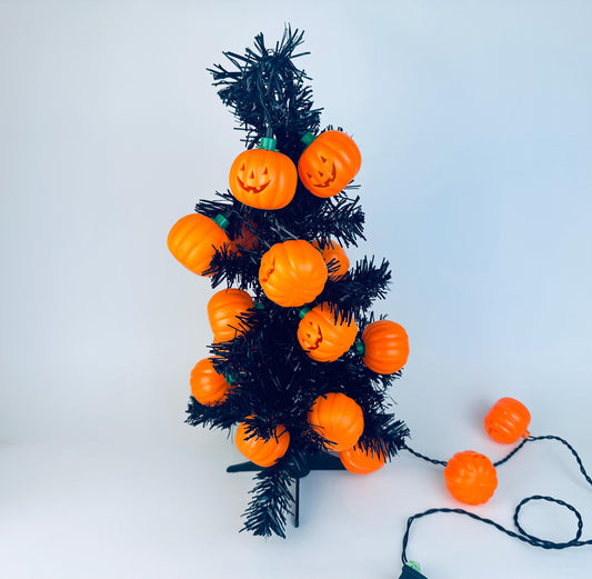 Black tabletop tree decorated with vintage orange plastic pumpkin jack-o’-lantern string lights, with extra pumpkins extending along the cord, displayed against a white background.