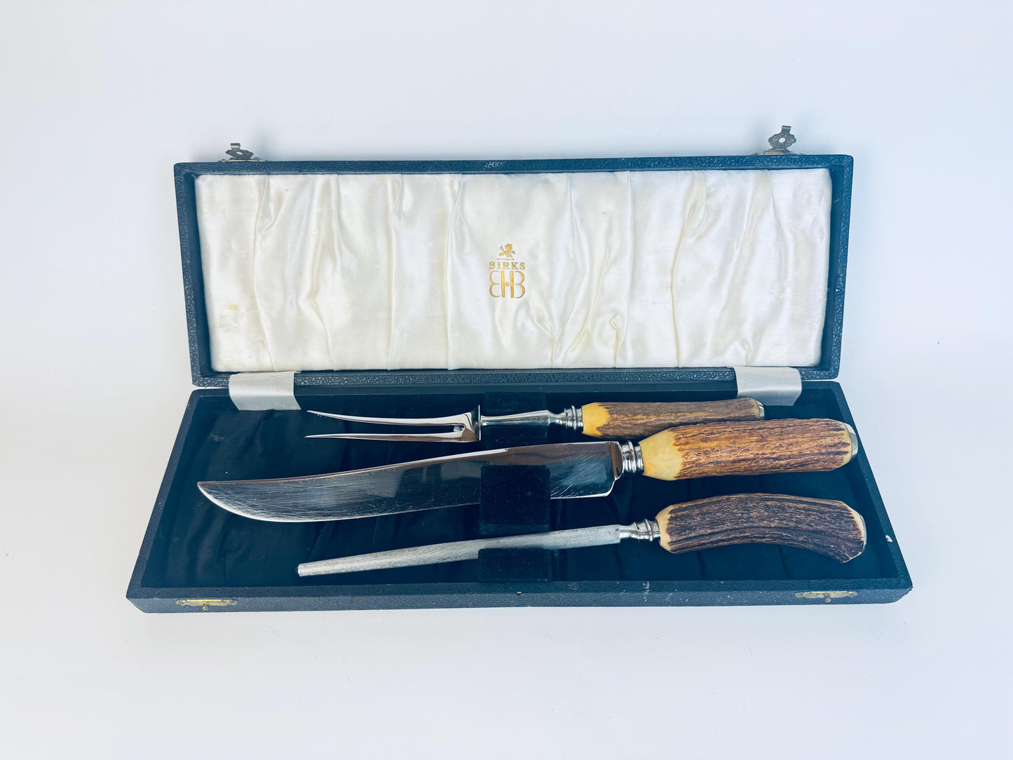 Vintage 1950s Birks three-piece carving set with stag-horn handles, stainless steel blades, and fork, displayed in its original black presentation case lined with ivory satin and marked “Birks” in gold lettering.