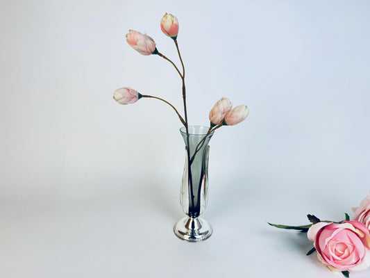 A slender vintage glass bud vase with a silver-tone base, holding several pale pink tulip stems against a soft white backdrop, with additional roses placed beside it for styling.