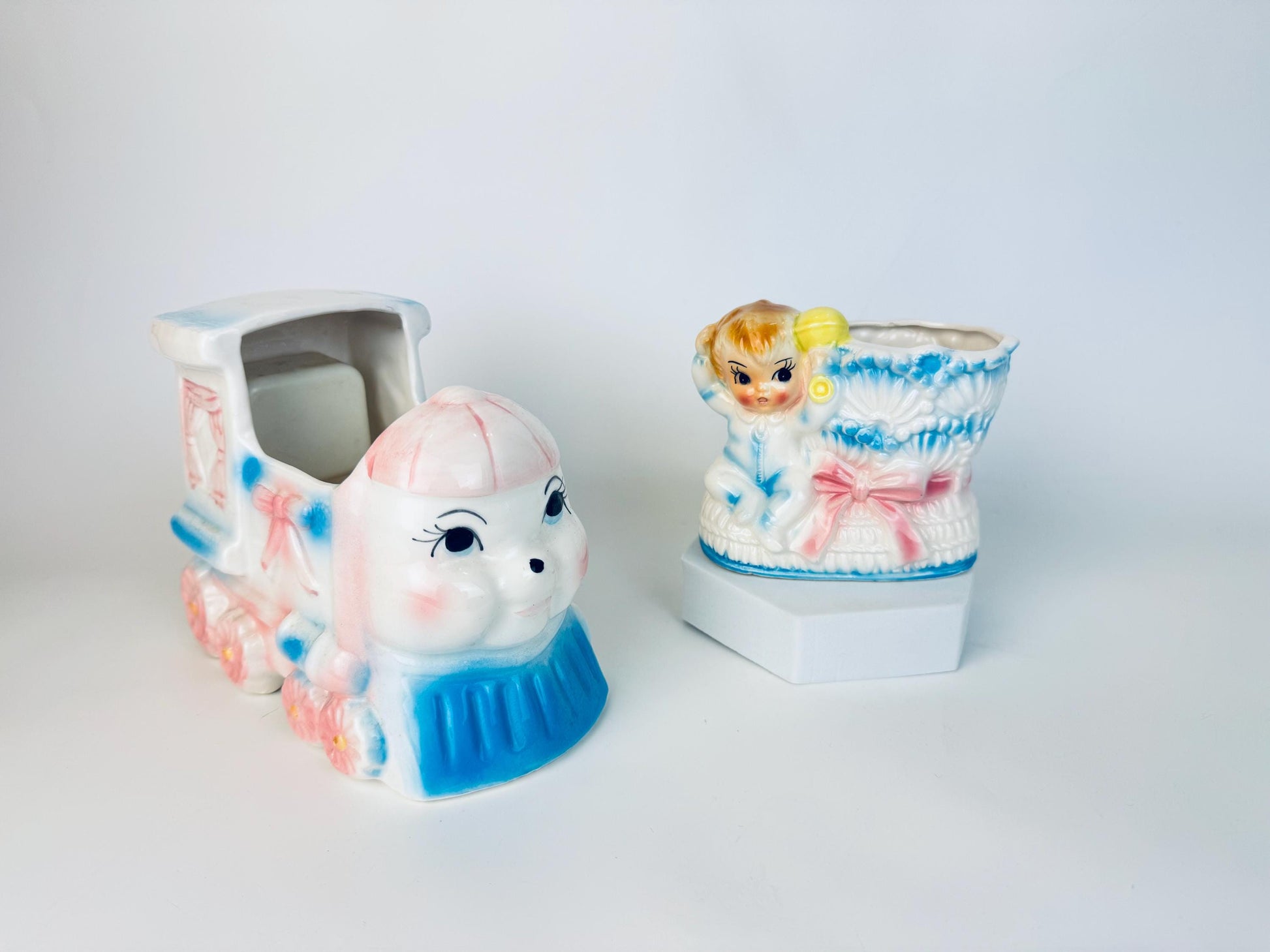 Pair of vintage 1950s–1960s ceramic nursery planters – one shaped like a smiling train engine in pastel pink and blue, the other a baby boot with a blond child in blue pyjamas climbing out, decorated with pink and blue bows.