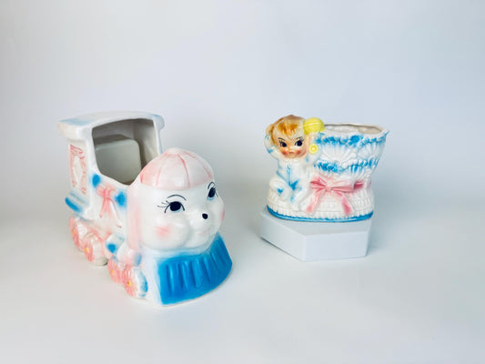 Pair of vintage 1950s–1960s ceramic nursery planters – one shaped like a smiling train engine in pastel pink and blue, the other a baby boot with a blond child in blue pyjamas climbing out, decorated with pink and blue bows.