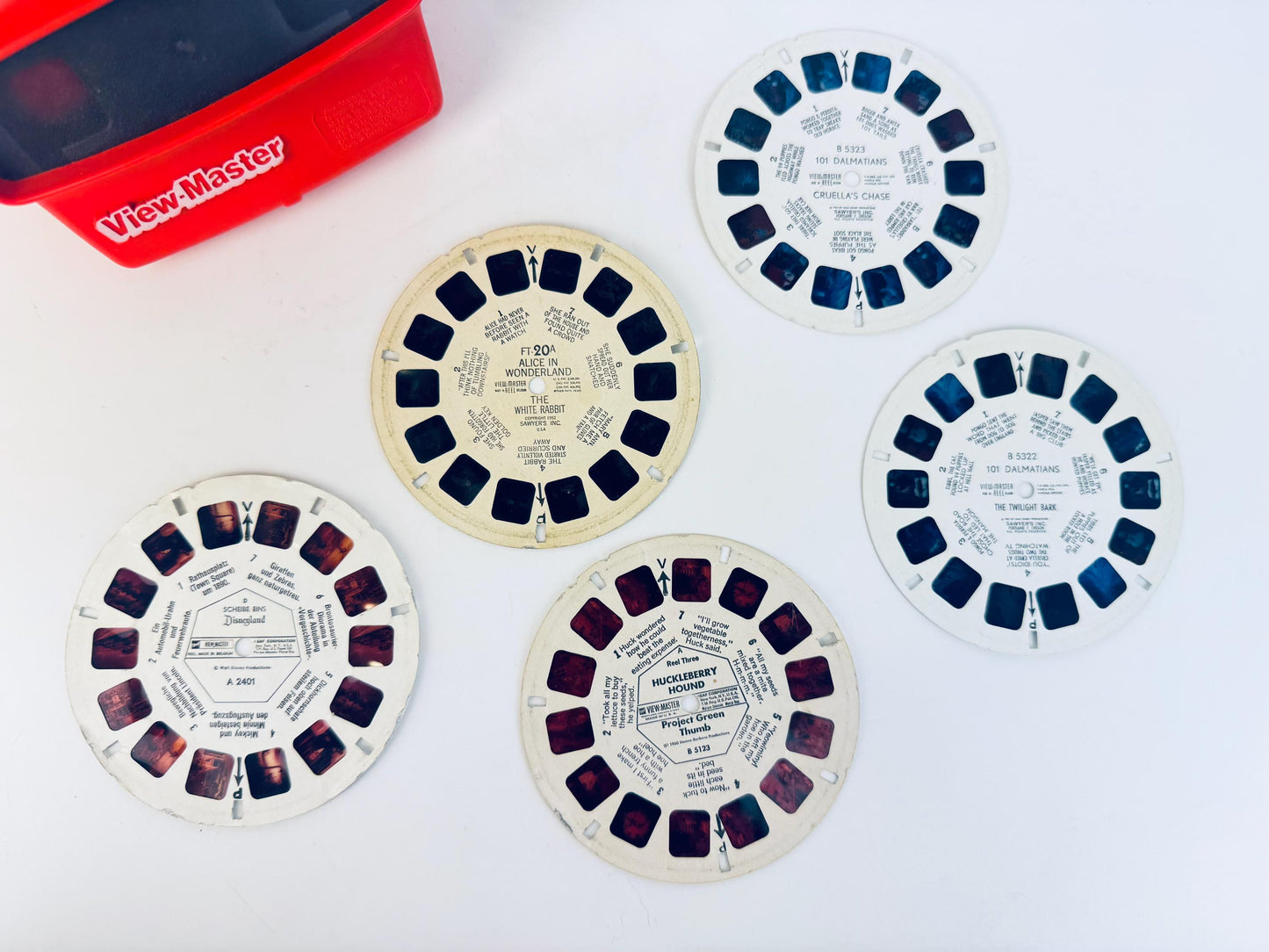 Vintage red View-Master with five stereoscopic reels laid out on a white surface, featuring titles including Alice in Wonderland, 101 Dalmatians, Huckberry Hound, and Disneyland scenes