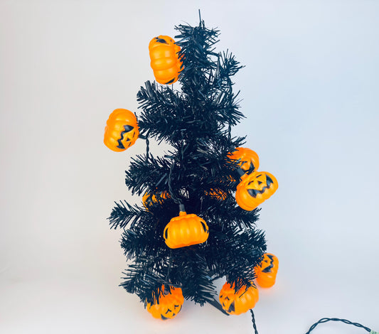 Small black artificial tree decorated with vintage orange plastic pumpkin jack-o’-lantern string lights, displayed on a white background.