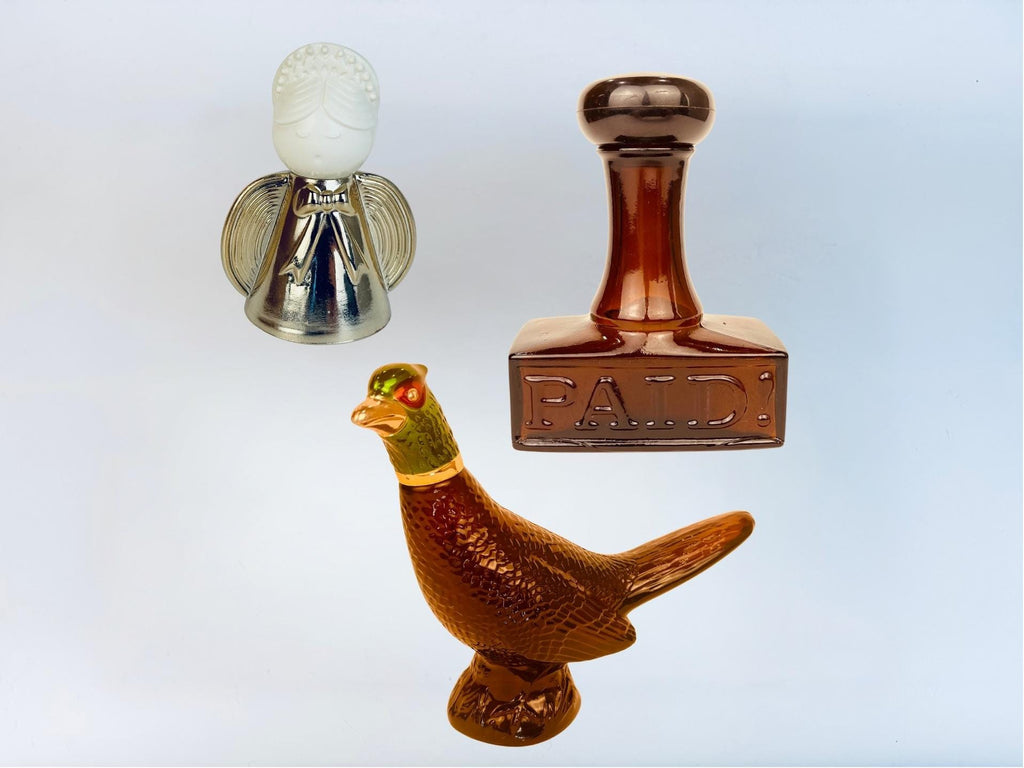 Three vintage Avon glass decanters from the 1970s, including a silver angel bottle, an amber “PAID” stamp bottle, and a brown and green pheasant-shaped bottle, displayed against a white background.