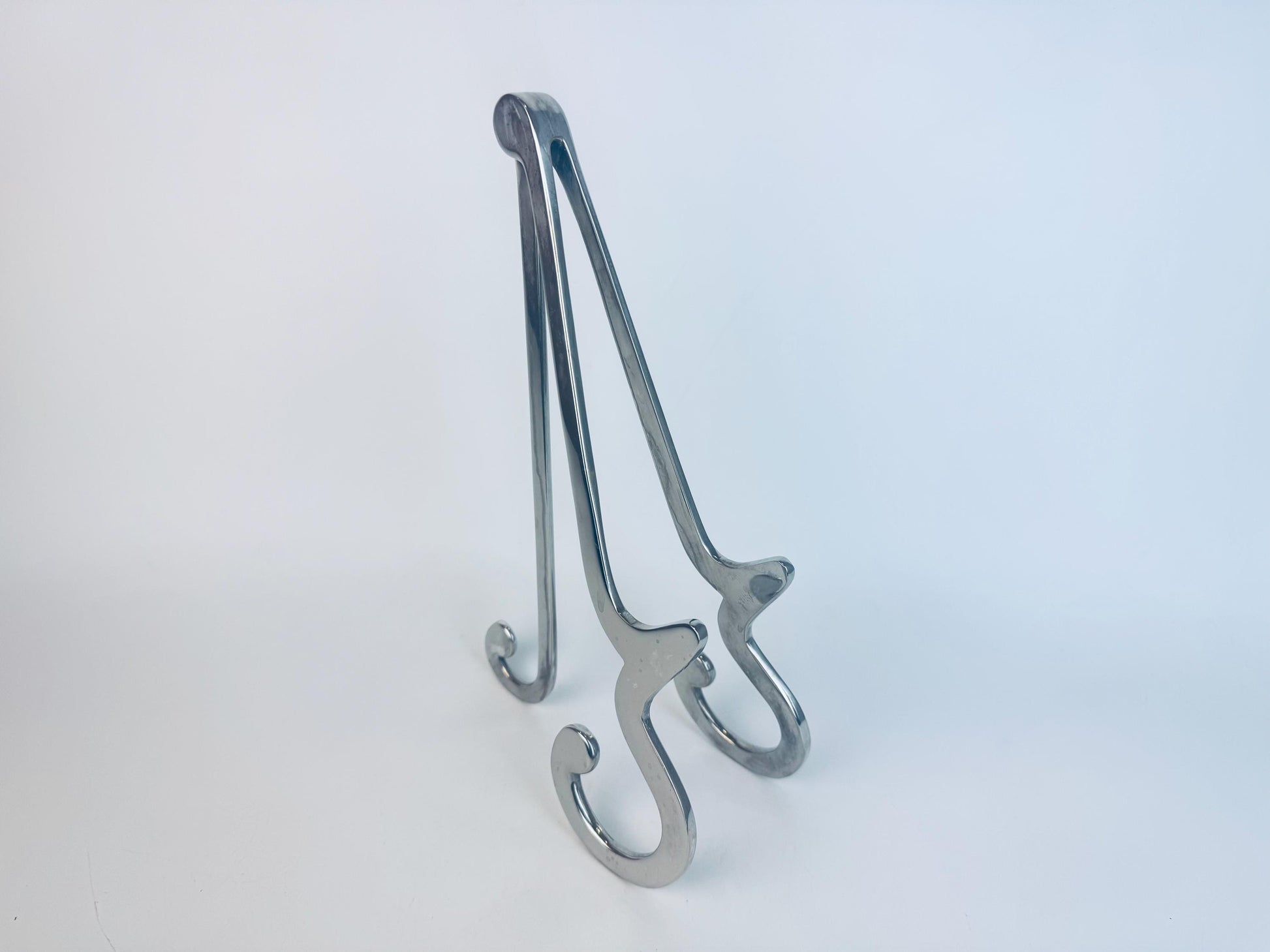 Polished chrome display easel with curved Art Deco–style feet and a minimalist folded-frame design, shown standing on a white background.