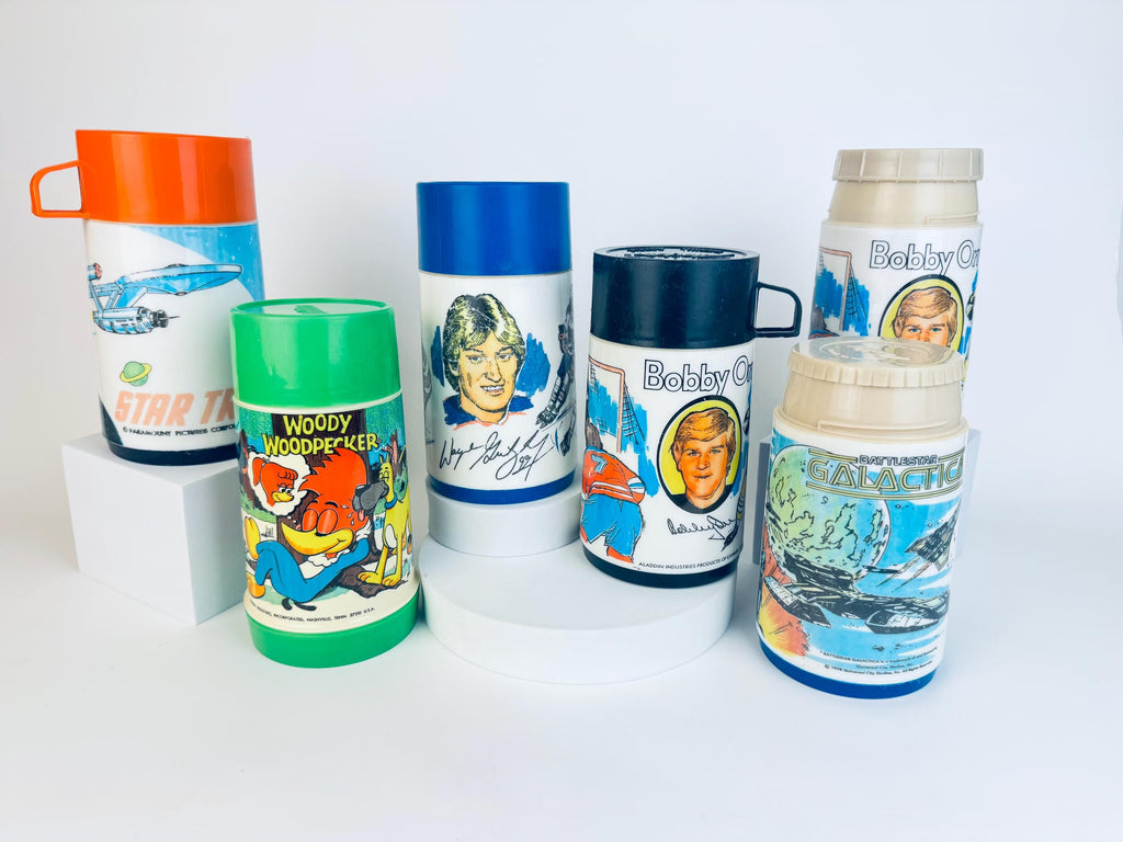 A collection of six vintage Aladdin thermoses from the 1970s and early 1980s, each featuring colourful graphics of popular characters and series. Designs include Star Trek, Woody Woodpecker, Wayne Gretzky, Bobby Orr, and Battlestar Galactica. The thermoses vary in lid colours—orange, green, blue, black, beige—and are displayed upright on white platforms against a clean white background.