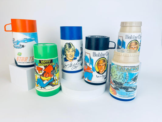 A collection of six vintage Aladdin thermoses from the 1970s and early 1980s, each featuring colourful graphics of popular characters and series. Designs include Star Trek, Woody Woodpecker, Wayne Gretzky, Bobby Orr, and Battlestar Galactica. The thermoses vary in lid colours—orange, green, blue, black, beige—and are displayed upright on white platforms against a clean white background.