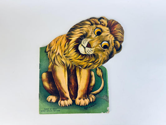 Vintage die-cut illustration of a lion with movable eyes and a tilting head, designed by artist Louis Wain. The lion is shown seated on a green background, with detailed fur and a curious, expressive face. The bottom corner includes patent information and the artist’s printed signature.