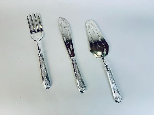Three vintage silver-toned serving utensils displayed on a white background — a fish fork, a fish knife, and a pie or cake server. Each piece features ornate engraved detailing on the handles and blades, showcasing an elegant floral and geometric motif.