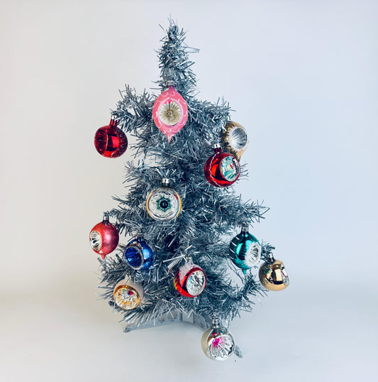 Small vintage silver tinsel Christmas tree decorated with colourful 1950s–1960s indented glass ornaments in red, pink, blue, gold, and green hues, displayed against a plain white background.