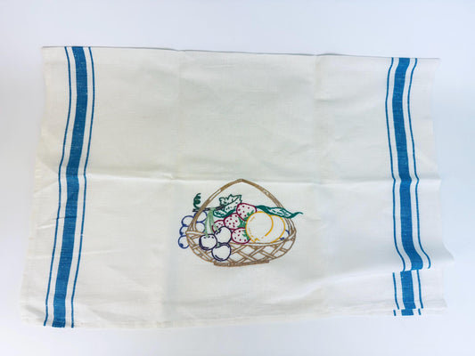 Vintage white linen tea towel featuring a woven basket filled with colourful fruit, framed by twin blue stripes along each side — classic farmhouse kitchen style.