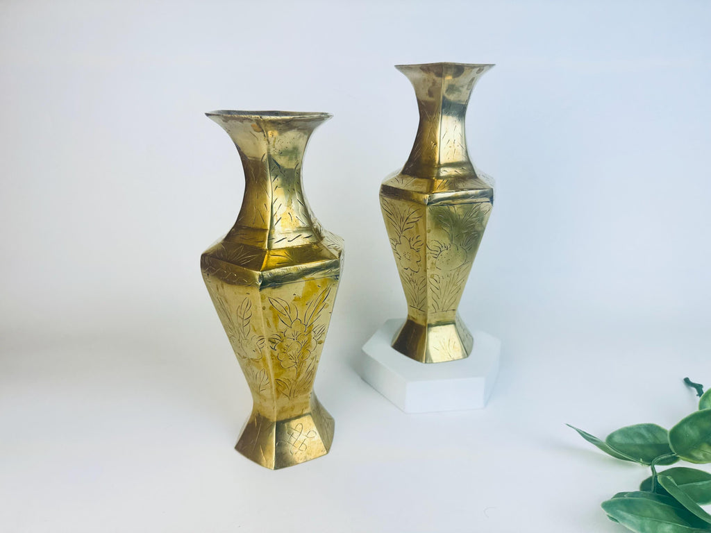 Two etched brass vases on a white background with a plant in the corner.