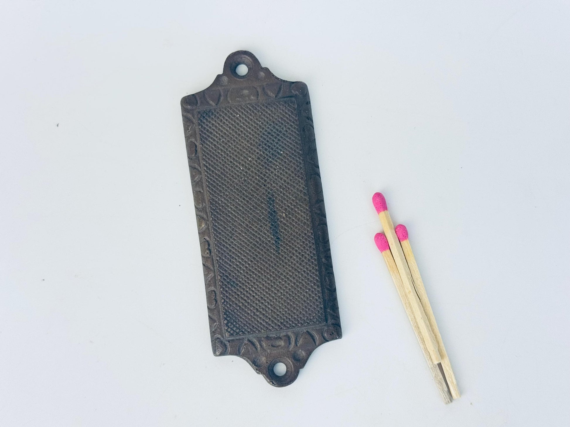 A vintage metal match striker plate with a textured surface and two mounting holes at each end, shown beside three wooden matches with pink tips for scale.