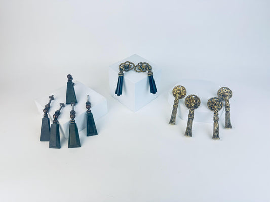 Three sets of vintage drawer pulls displayed on white blocks — on the left, a group of dark geometric Bakelite pulls with metal fittings; in the centre, a pair of black and brass Art Deco-style tassel pulls; and on the right, four ornate brass tassel pulls with intricate circular backplates.