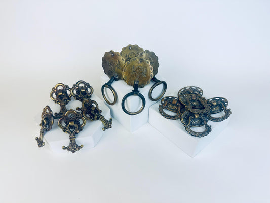 Three sets of ornate vintage brass drawer pulls arranged on white blocks — on the left, a group of Rococo-style keyhole pulls with scrollwork designs; in the centre, round scalloped pulls engraved with floral patterns and ring handles; and on the right, intricately detailed circular pulls with openwork edging and raised centre motifs.