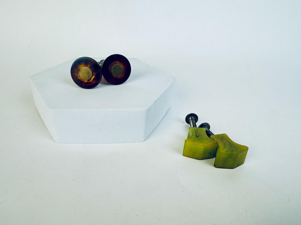Two pairs of vintage Bakelite drawer pulls displayed on a white surface — one pair in a round amber-brown tortoiseshell design with brass centres, and another pair in a sculpted green swirl with metal screws attached.