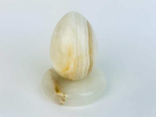 Vintage 1970s honey-banded ivory onyx egg displayed upright on its matching rounded stone stand, showing natural golden veining and a smooth polished finish.