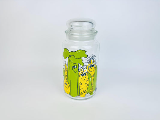 Vintage 1970s clear glass canister jar with a fitted glass lid, featuring Hildi’s whimsical green celery and yellow corn characters printed on the front.