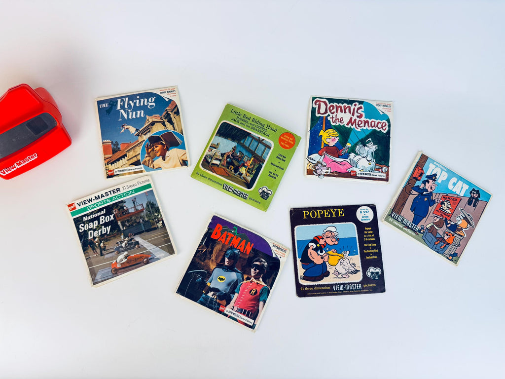 Vintage 1960s View-Master reel collection displayed on a white surface, including The Flying Nun, Little Red Riding Hood trio storybook reel, Dennis the Menace, Top Cat, National Soap Box Derby, Batman, and Popeye packets with a red View-Master viewer on the left.