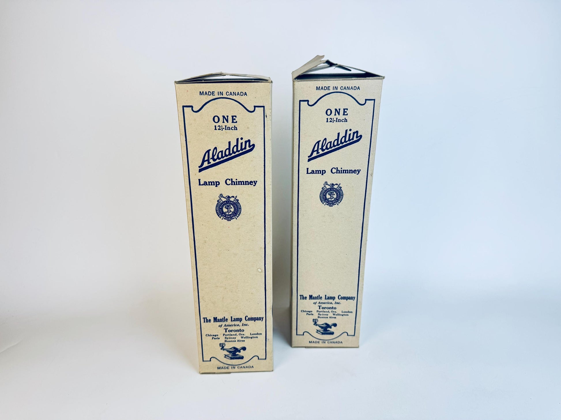 Two vintage Aladdin 12½-inch lamp chimney boxes made in Canada by The Mantle Lamp Company of America, Toronto. The beige packaging features blue lettering and graphics, including the Aladdin logo and an illustration of an oil lamp, displayed upright against a white background.