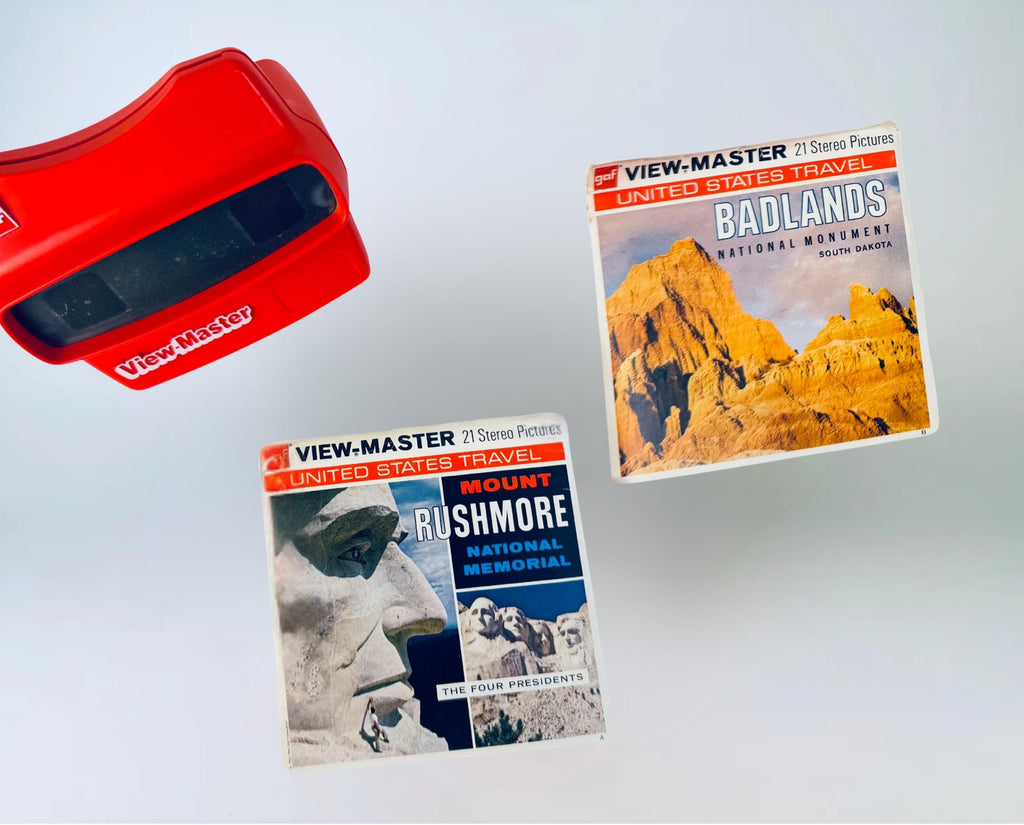 Red vintage View-Master viewer displayed with two GAF View-Master reel packets—‘Badlands National Monument’ and ‘Mount Rushmore National Memorial,’ both from the United States Travel series—set against a soft grey background.