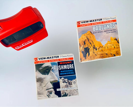 Red vintage View-Master viewer displayed with two GAF View-Master reel packets—‘Badlands National Monument’ and ‘Mount Rushmore National Memorial,’ both from the United States Travel series—set against a soft grey background.