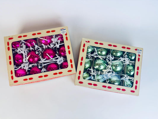 Two boxes of vintage 1960s Coby mercury-glass Christmas ornaments, one set in bright fuchsia pink and the other in mint green, shown in their original cream boxes with red holly borders and clear window lids.