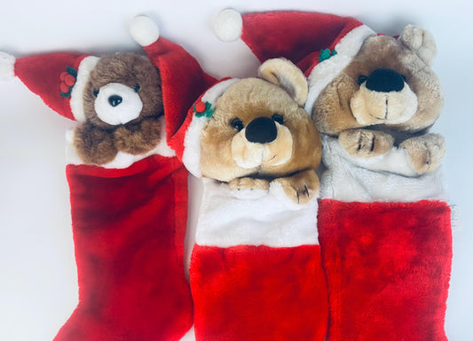 Three vintage plush bear Christmas stockings arranged side by side, each with a soft red stocking body and a teddy bear head wearing a red Santa hat with holly detail. The bears vary in colour from dark brown to tan to light beige, all with their plush paws resting over the stocking edge.