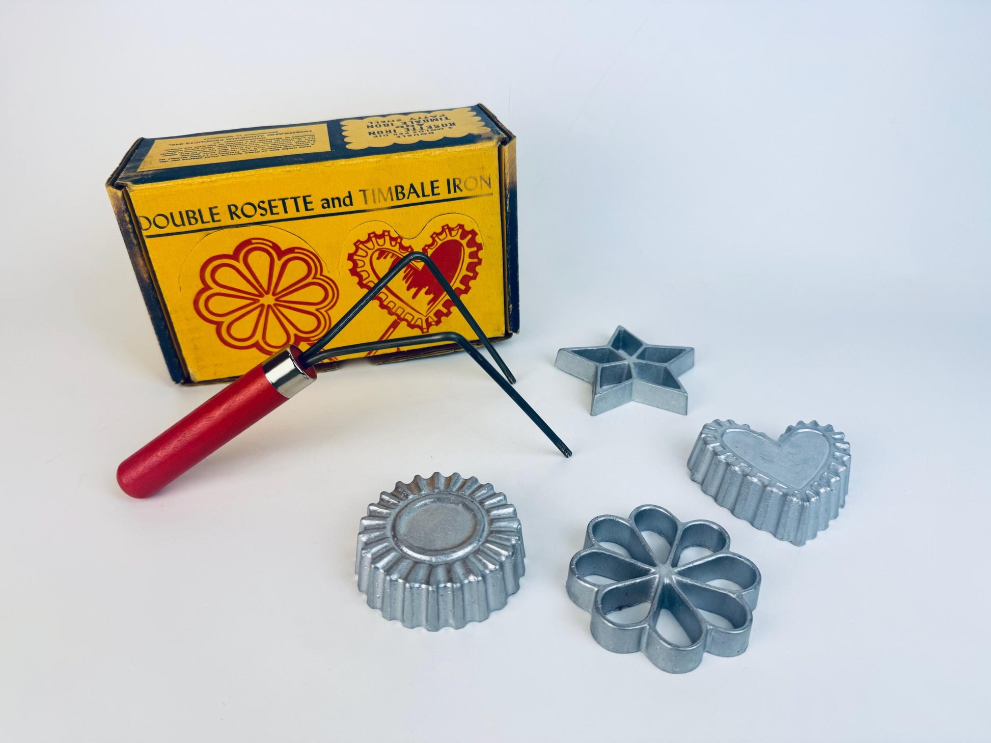 Vintage Nordic Ware rosette and timbale iron set displayed with the original yellow box, red-handled iron, and four silver cast-aluminium moulds (star, heart, flower, and scalloped cup) arranged on a white background.