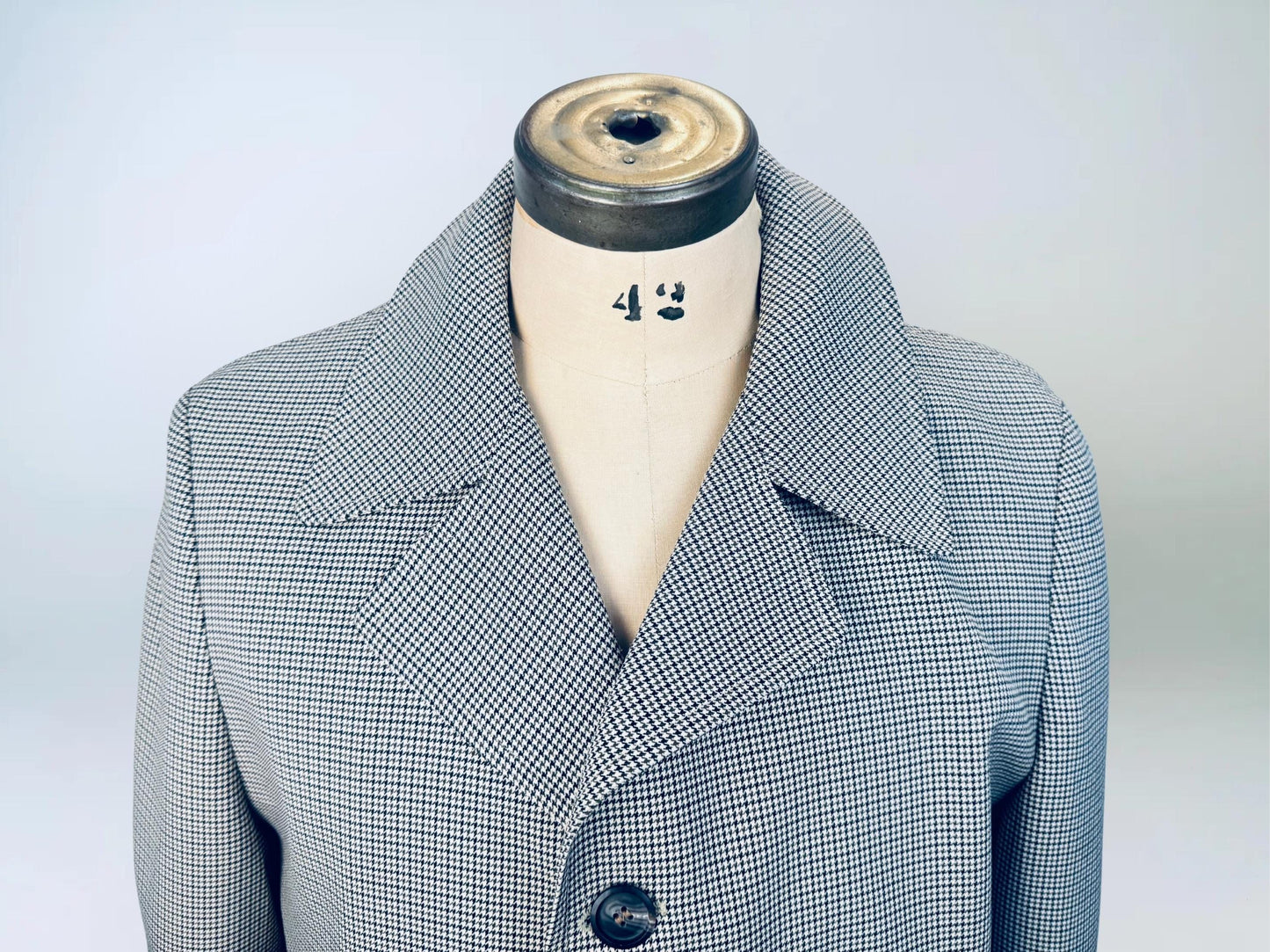 Close-up of a vintage men’s trench coat displayed on a dress form, featuring a fine black-and-brown houndstooth pattern and wide notched lapel collar.