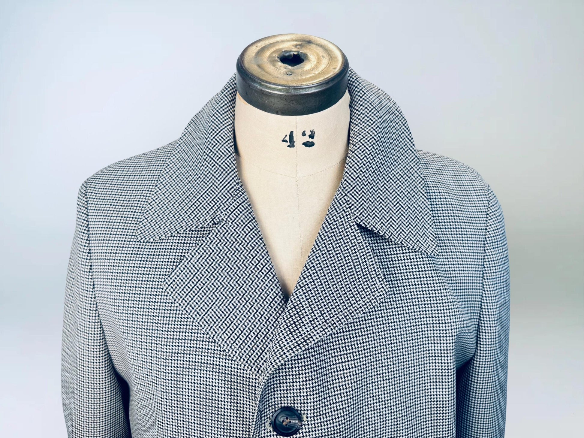 Close-up of a vintage men’s trench coat displayed on a dress form, featuring a fine black-and-brown houndstooth pattern and wide notched lapel collar.