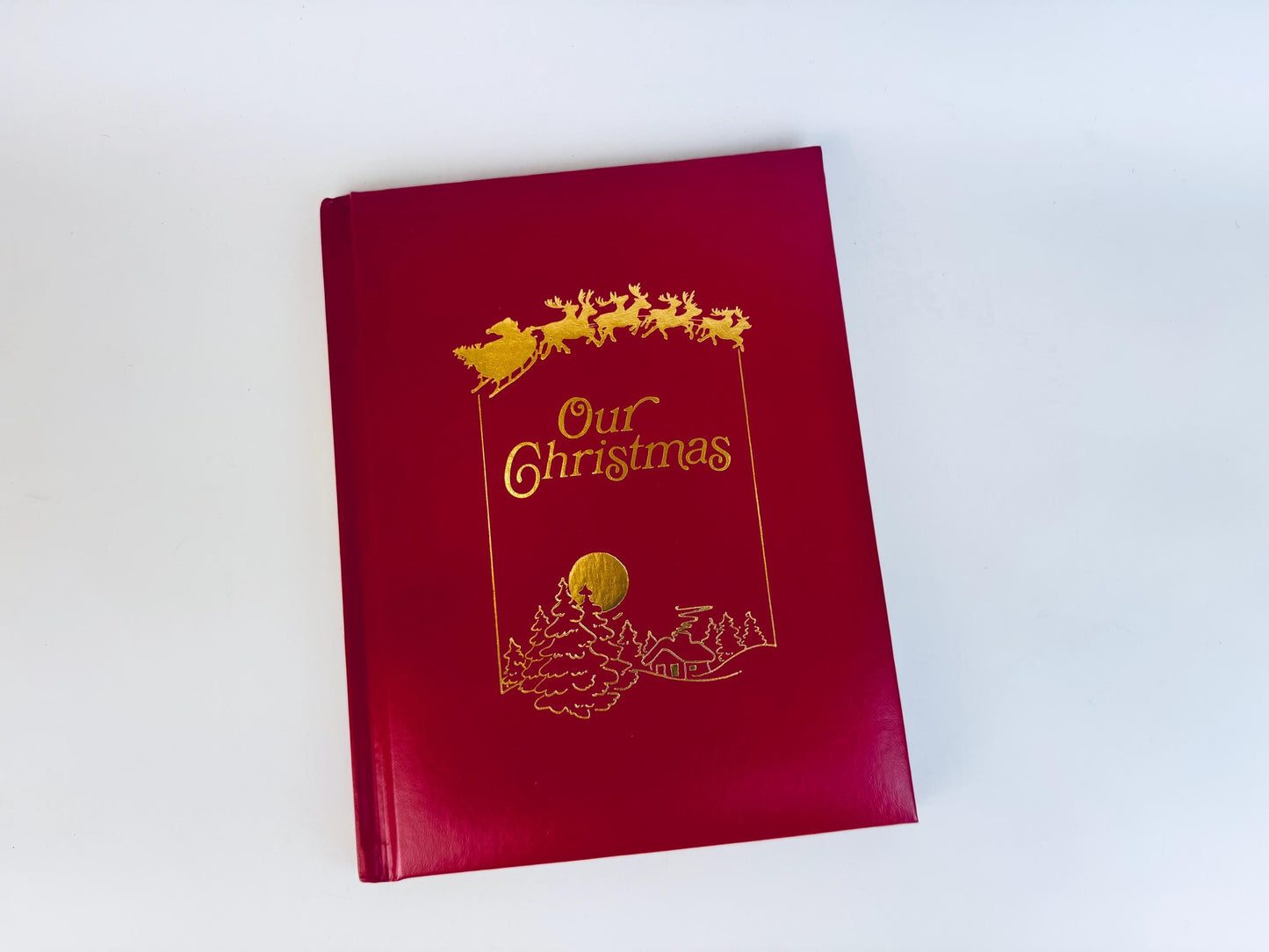 Vintage red leatherette hardcover book titled ‘Our Christmas’ with gold foil sleigh and reindeer illustration on the front cover.