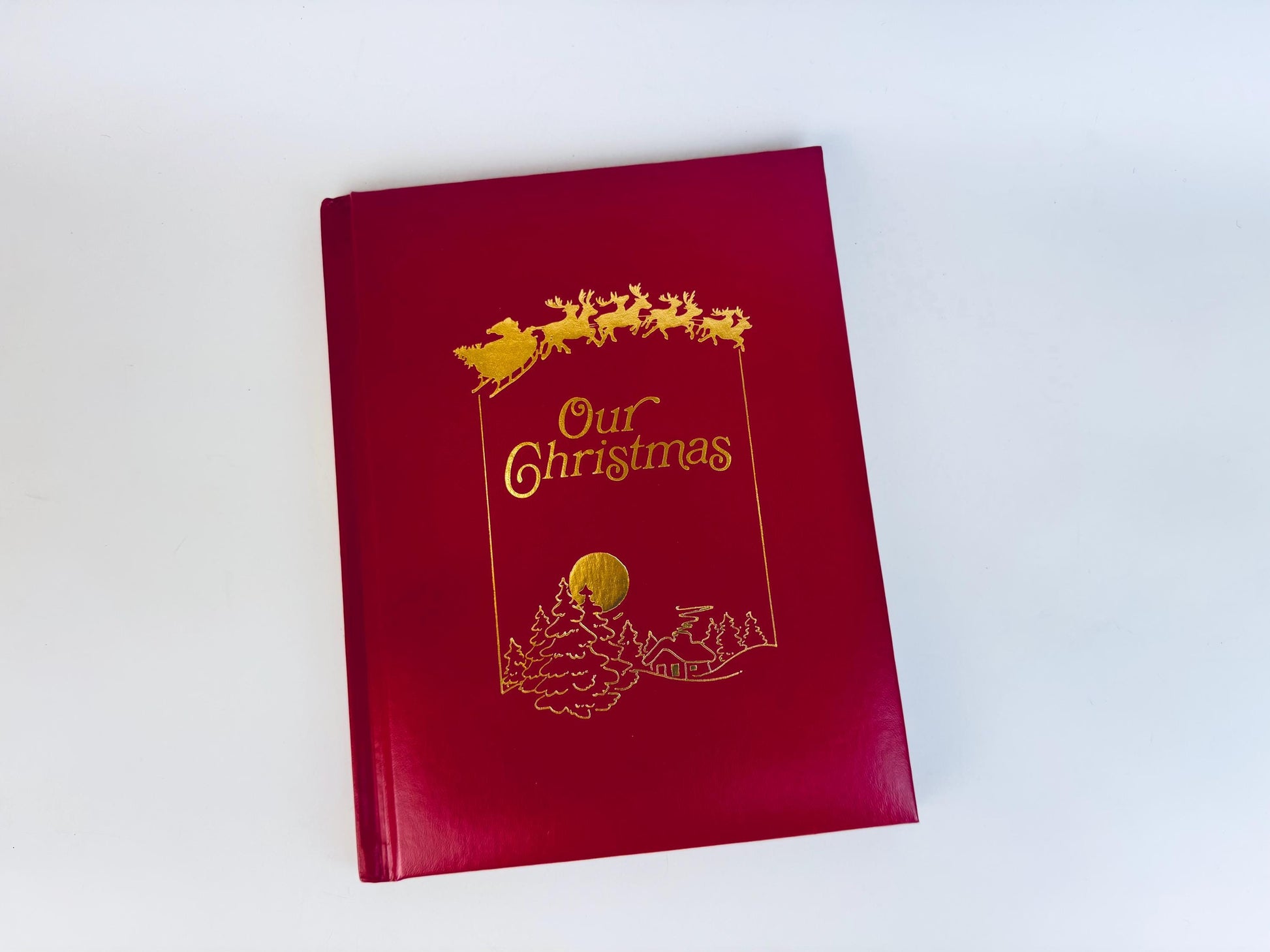 Vintage red leatherette hardcover book titled ‘Our Christmas’ with gold foil sleigh and reindeer illustration on the front cover.