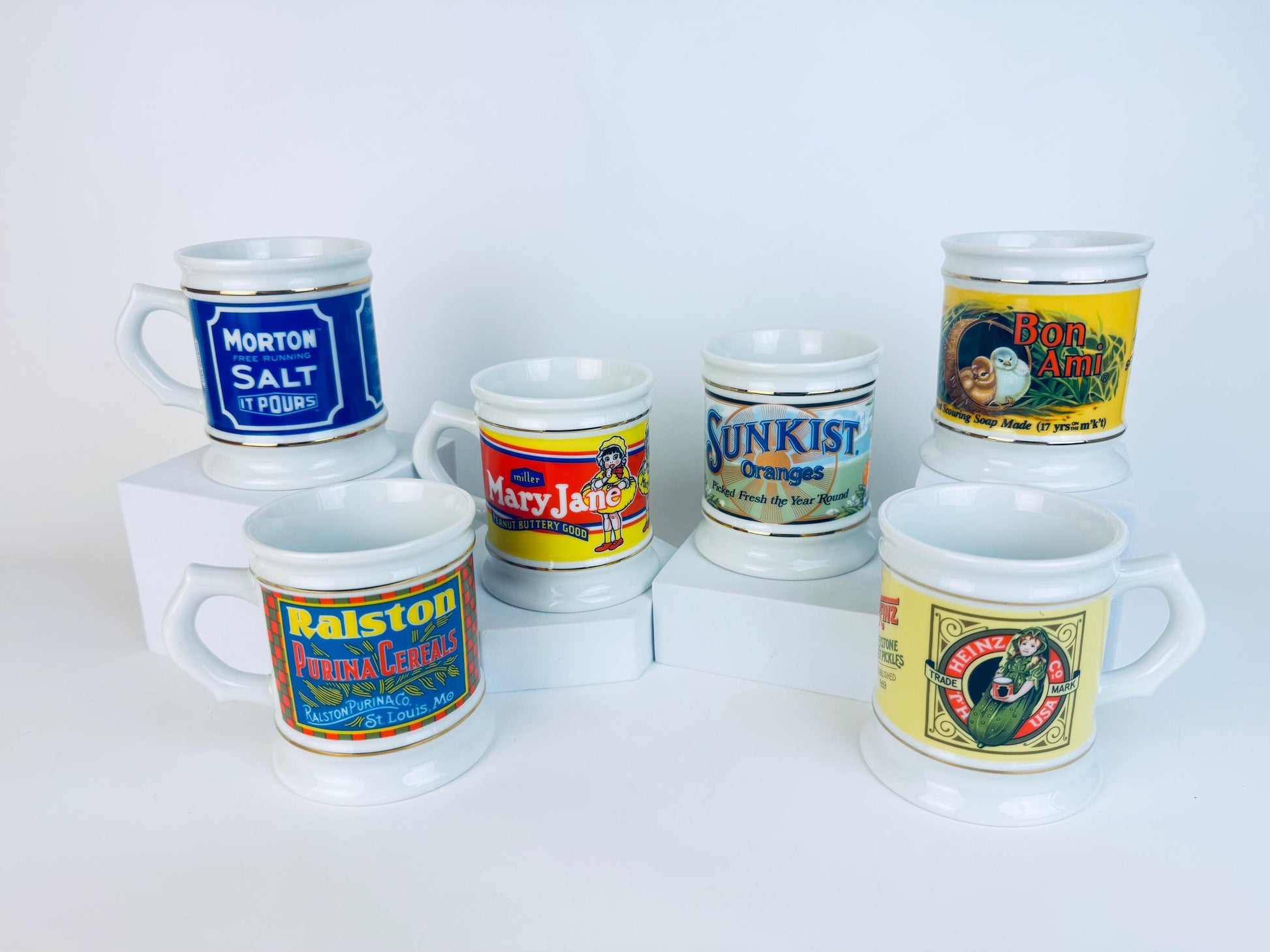 Group of six 1984 Franklin Porcelain advertising mugs displayed on white blocks, each sold individually with colourful vintage brand graphics and gold-trimmed white porcelain bodies.