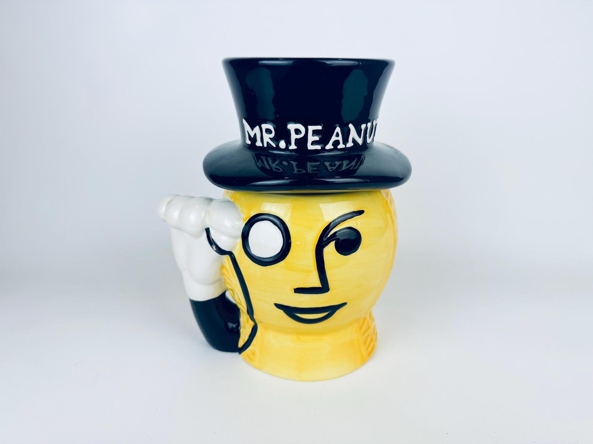Figural ceramic cookie jar in the shape of Mr Peanut wearing a black top hat, glossy yellow body textured like a peanut shell.
