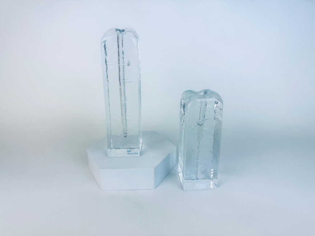 Pair of vintage 1970s Muurla Finland clear ice-glass candleholders, tall and short column shapes with vertical inner channels, displayed on a white geometric stand.