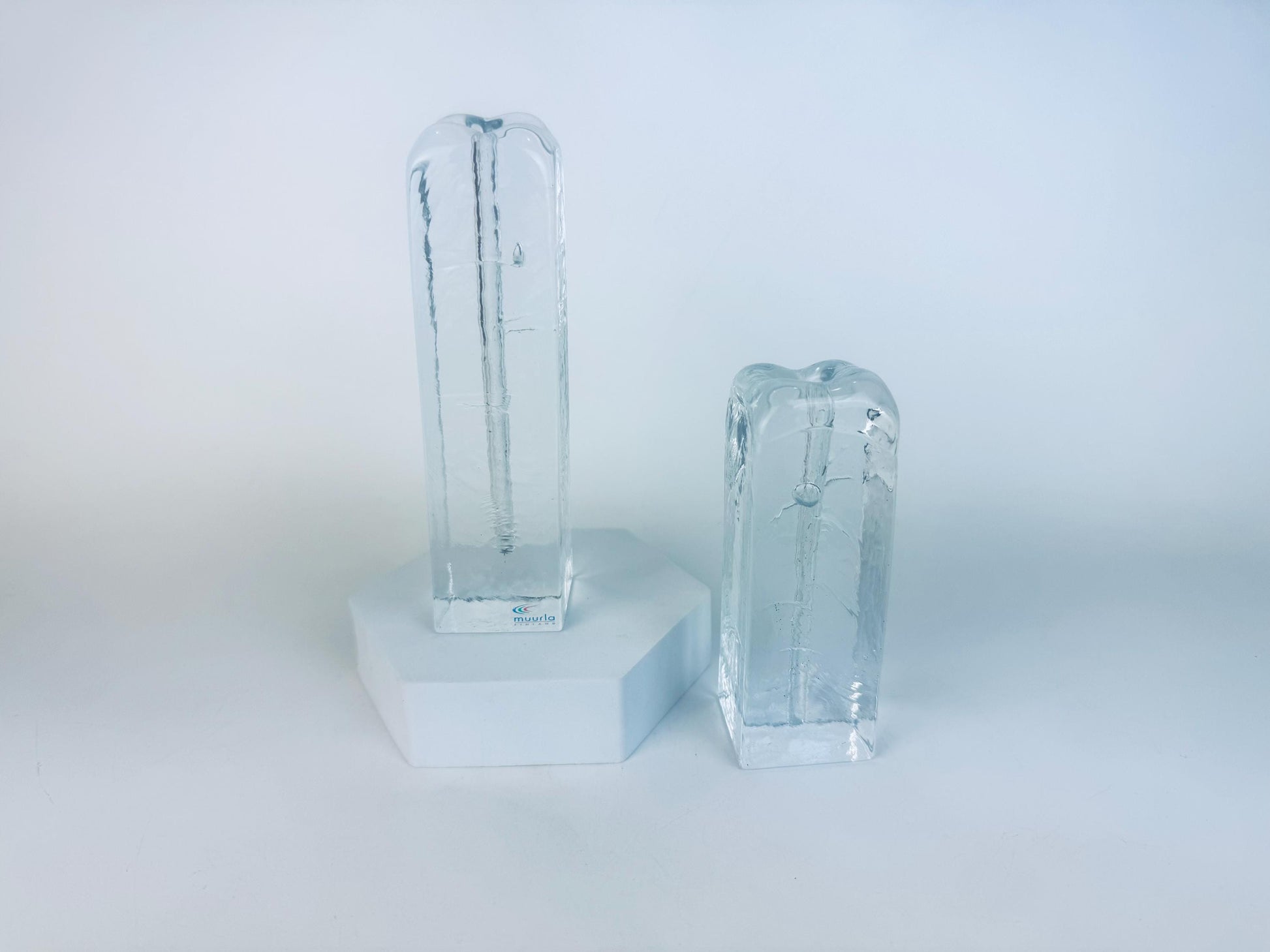 Pair of vintage 1970s Muurla Finland clear ice-glass candleholders, tall and short column shapes with vertical inner channels, displayed on a white geometric stand.