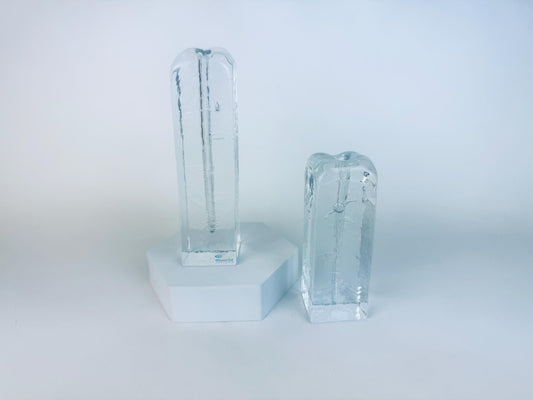 Pair of vintage 1970s Muurla Finland clear ice-glass candleholders, tall and short column shapes with vertical inner channels, displayed on a white geometric stand.
