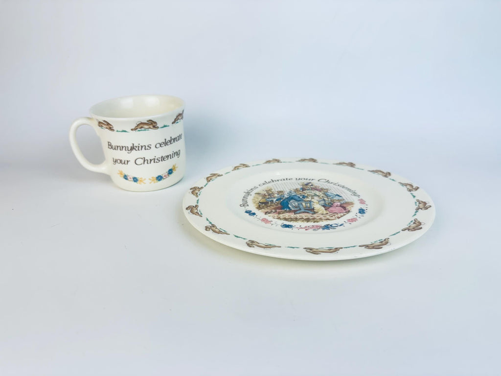 Vintage Royal Doulton Bunnykins Christening set featuring a cream bone china mug and matching plate with running rabbit borders and a colourful christening scene, displayed on a white background.