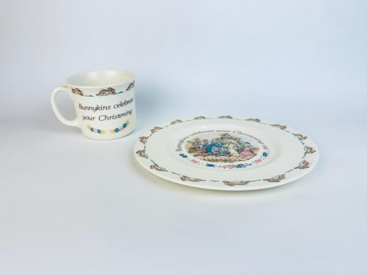 Vintage Royal Doulton Bunnykins Christening set featuring a cream bone china mug and matching plate with running rabbit borders and a colourful christening scene, displayed on a white background.