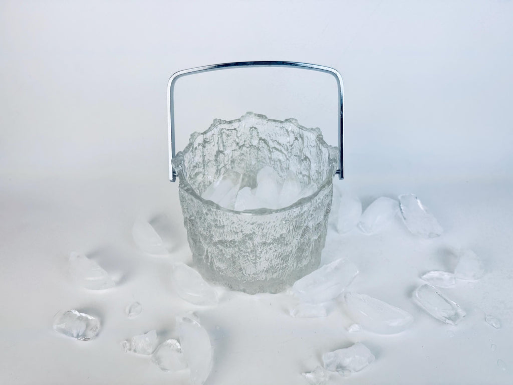 Vintage 1970s Pukeberg Sweden clear textured glass ice bucket with chrome handle, surrounded by scattered ice pieces on a white background.