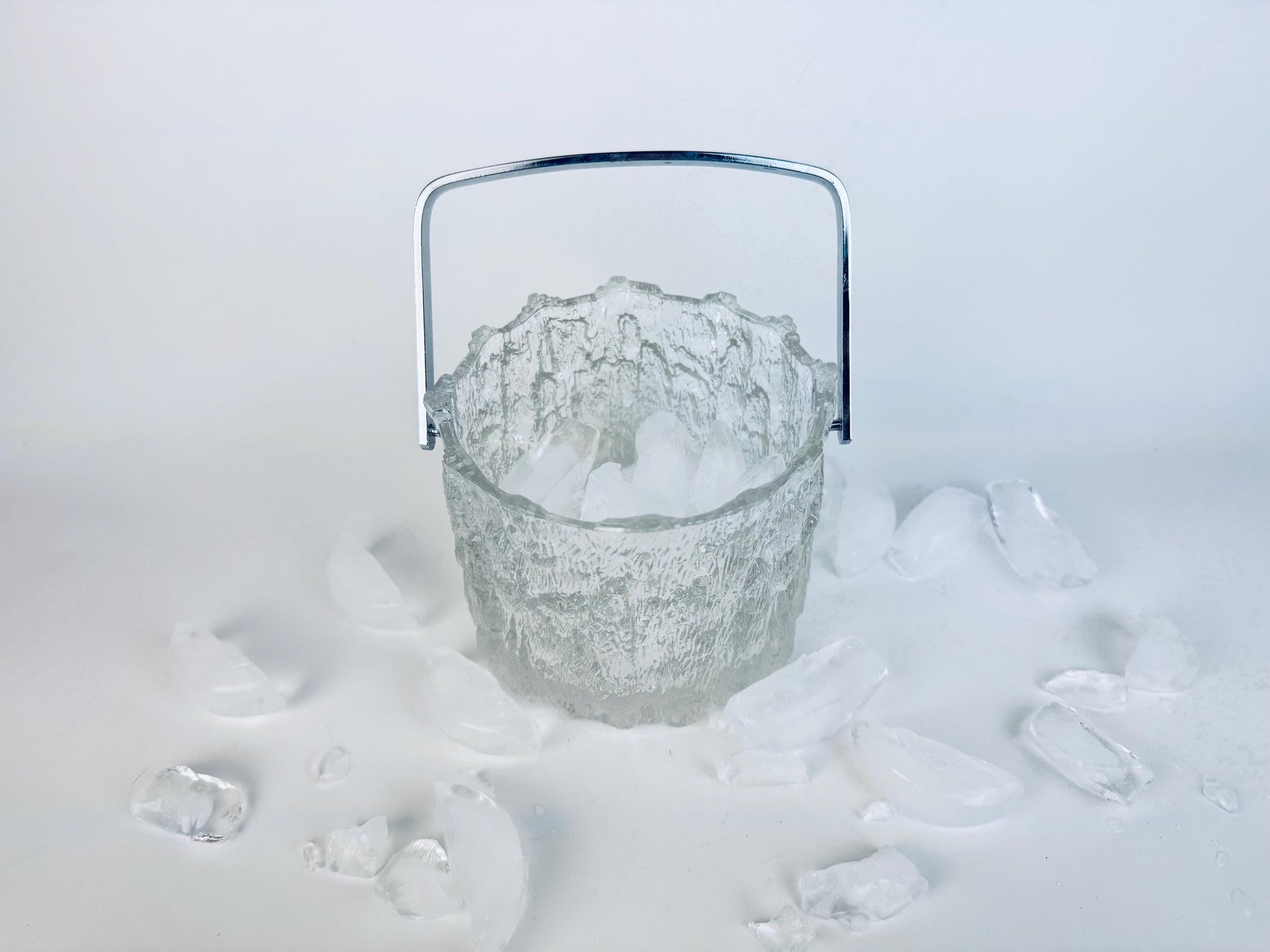 Vintage 1970s Pukeberg Sweden clear textured glass ice bucket with chrome handle, surrounded by scattered ice pieces on a white background.