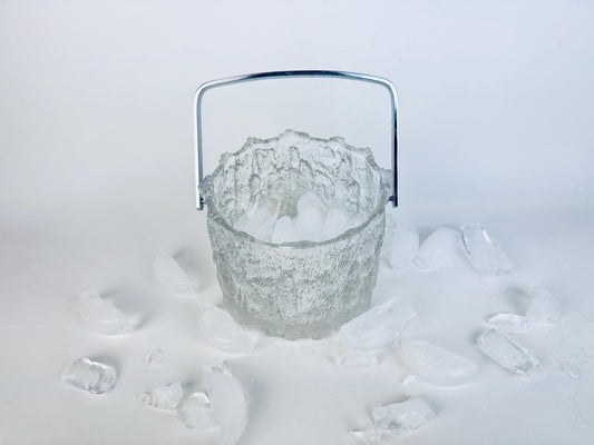 Vintage 1970s Pukeberg Sweden clear textured glass ice bucket with chrome handle, surrounded by scattered ice pieces on a white background.