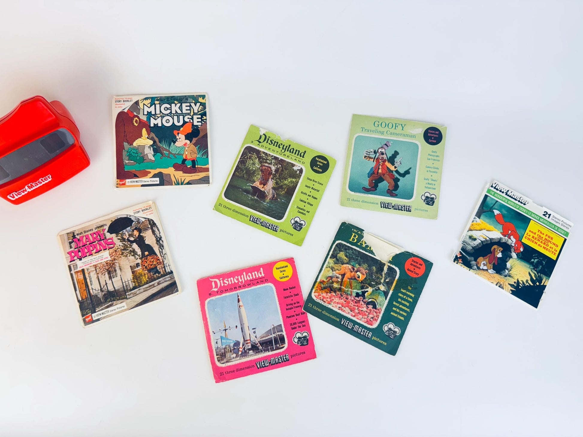 Vintage Disney View-Master reel collection displayed on a white surface, including Mickey Mouse, Goofy Traveling Cameraman, Bambi, The Fox and the Hound, Mary Poppins, Disneyland Adventureland, and Disneyland Tomorrowland packets with a red View-Master viewer on the left.