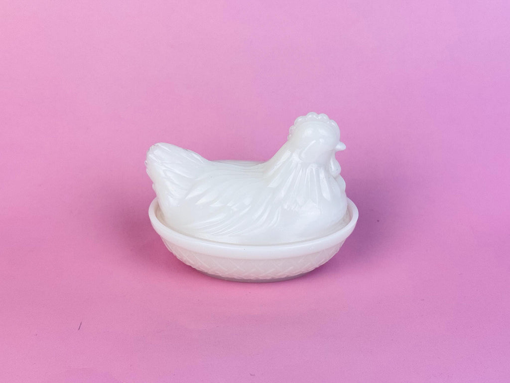 White vintage milk-glass hen-on-nest dish shown against a soft pink background, featuring a smooth opaline finish, detailed feather moulding, and a crosshatch-patterned base.