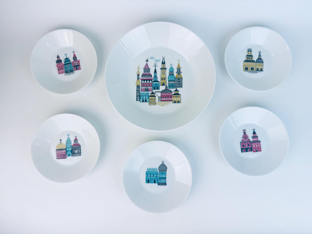 Overhead view of a six-piece vintage German porcelain bowl set featuring colourful illustrated city-scene buildings in teal, magenta, and mustard on white glaze, arranged neatly on a white background.